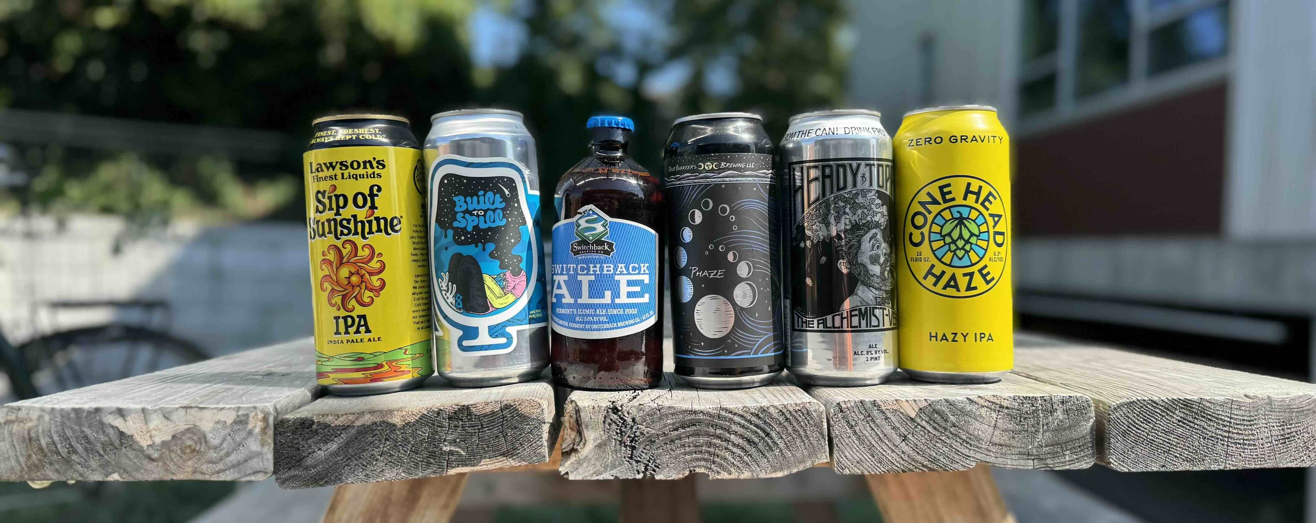6 VT beers aligned in a row on top of a picnic table. From left to right: Lawson's Sip of Sunshine, Foam Built to Spill IPA, Switchback Ale, Four Quarters Phaze IPA, Alchemist Heady Topper IPA, and Zero Gravity Conehead Haze IPA. All are brewed in Vermont