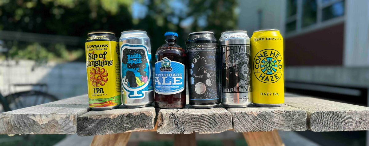 6 VT beers aligned in a row on top of a picnic table. From left to right: Lawson's Sip of Sunshine, Foam Built to Spill IPA, Switchback Ale, Four Quarters Phaze IPA, Alchemist Heady Topper IPA, and Zero Gravity Conehead Haze IPA. All are brewed in Vermont